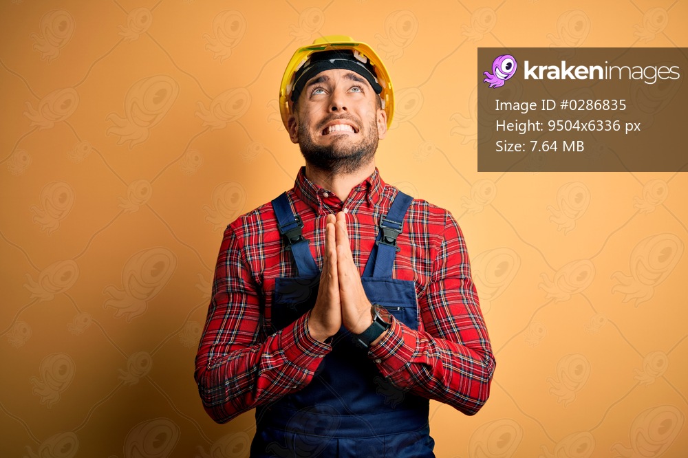 Young builder man wearing construction uniform and safety helmet over yellow isolated background begging and praying with hands together with hope expression on face very emotional and worried. Begging.