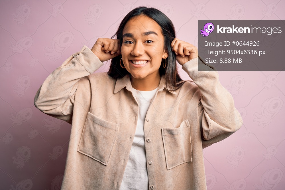 Young beautiful asian woman wearing casual shirt standing over pink background Smiling pulling ears with fingers, funny gesture. Audition problem