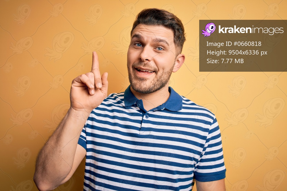 Young man with blue eyes wearing casual striped t-shirt over yellow background pointing finger up with successful idea. Exited and happy. Number one.