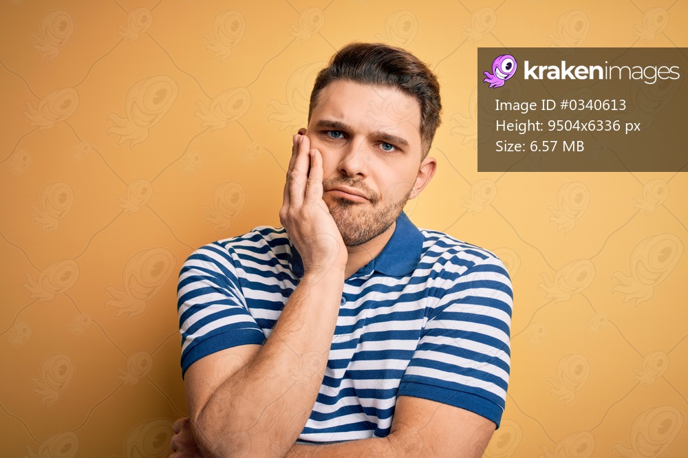Young man with blue eyes wearing casual striped t-shirt over yellow background thinking looking tired and bored with depression problems with crossed arms.