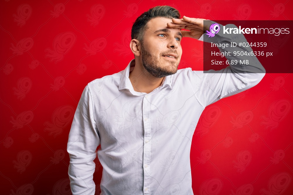 Young business man with blue eyes wearing elegant shirt standing over red isolated background very happy and smiling looking far away with hand over head. Searching concept.