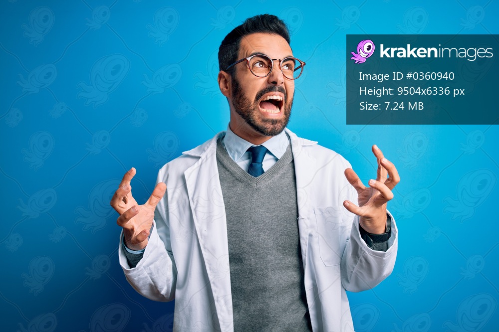 Young handsome doctor man with beard wearing coat and glasses over blue background crazy and mad shouting and yelling with aggressive expression and arms raised. Frustration concept.