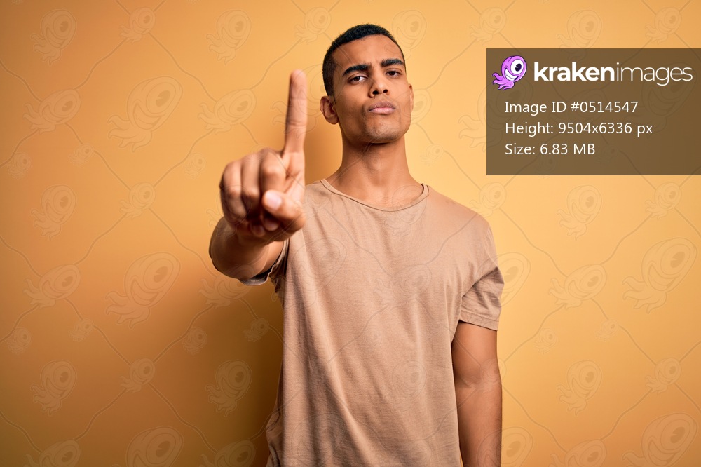 Young handsome african american man wearing casual t-shirt standing over yellow background Pointing with finger up and angry expression, showing no gesture