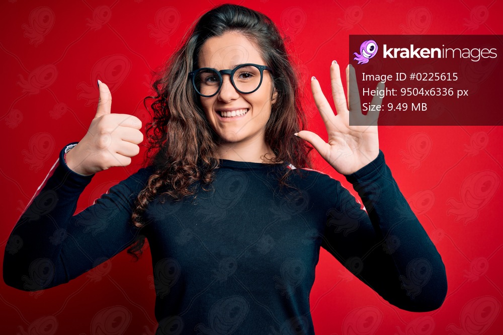 Young beautiful woman with curly hair wearing sweater and glasses over red background showing and pointing up with fingers number six while smiling confident and happy.