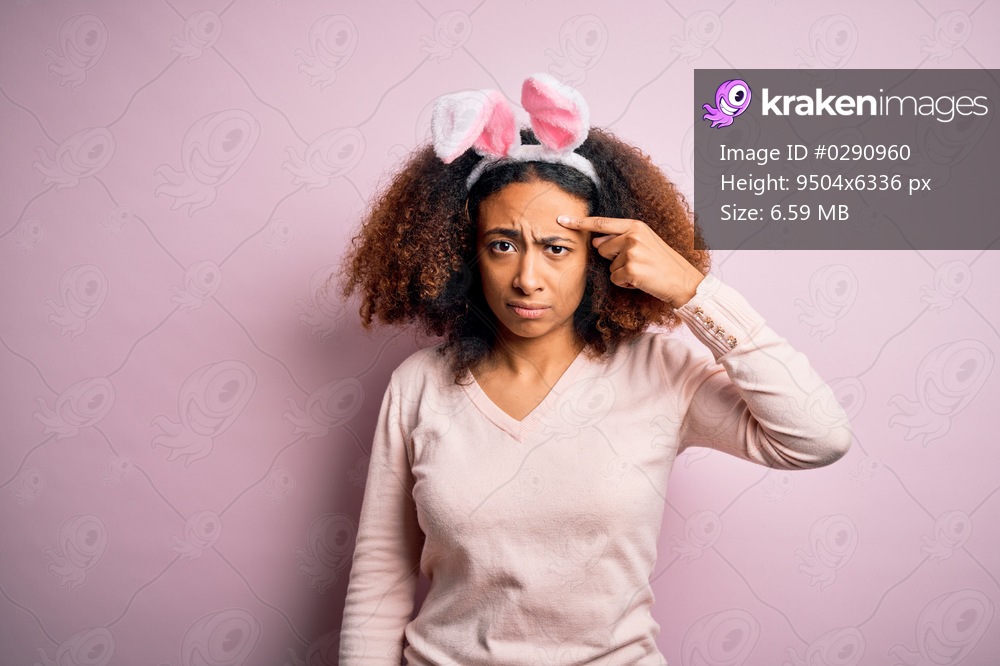 Young african american woman with afro hair wearing bunny ears over pink background pointing unhappy to pimple on forehead, ugly infection of blackhead. Acne and skin problem