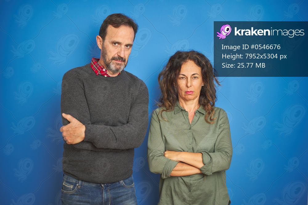 Beautiful middle age couple together standing over isolated blue background skeptic and nervous, disapproving expression on face with crossed arms. Negative person.