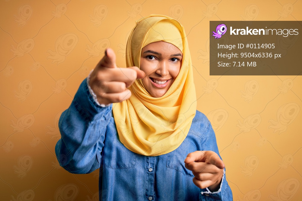 Young beautiful woman with curly hair wearing arab traditional hijab over yellow background pointing to you and the camera with fingers, smiling positive and cheerful