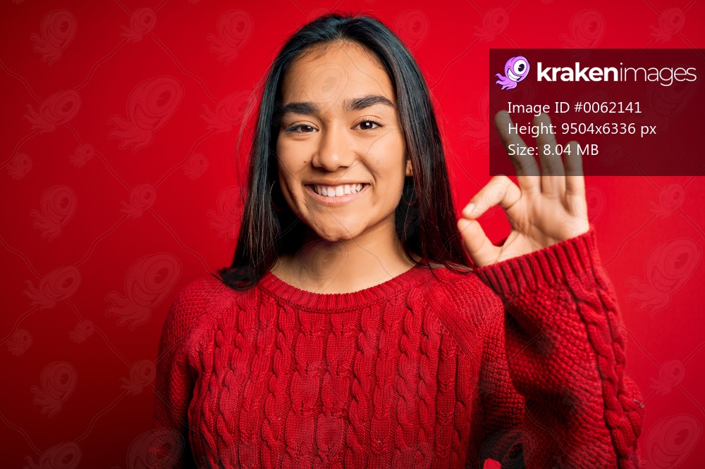 Young beautiful asian woman wearing casual sweater standing over isolated red background smiling positive doing ok sign with hand and fingers. Successful expression.
