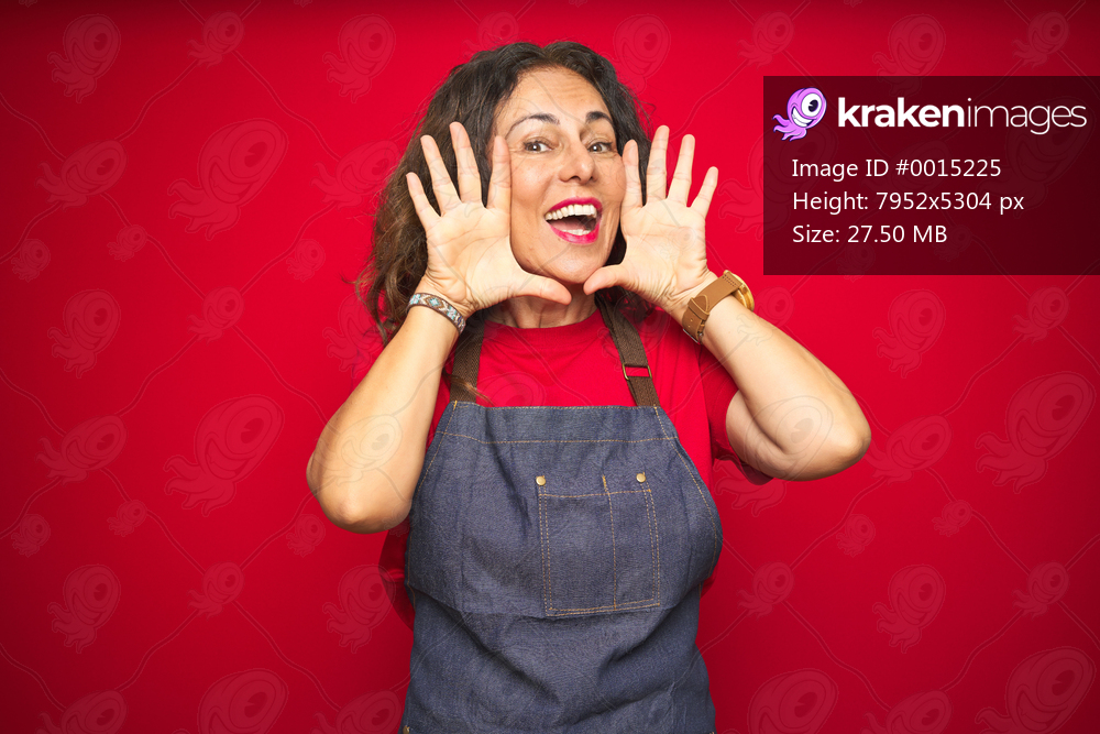 Middle age senior woman wearing apron uniform over red isolated background Smiling cheerful playing peek a boo with hands showing face. Surprised and exited