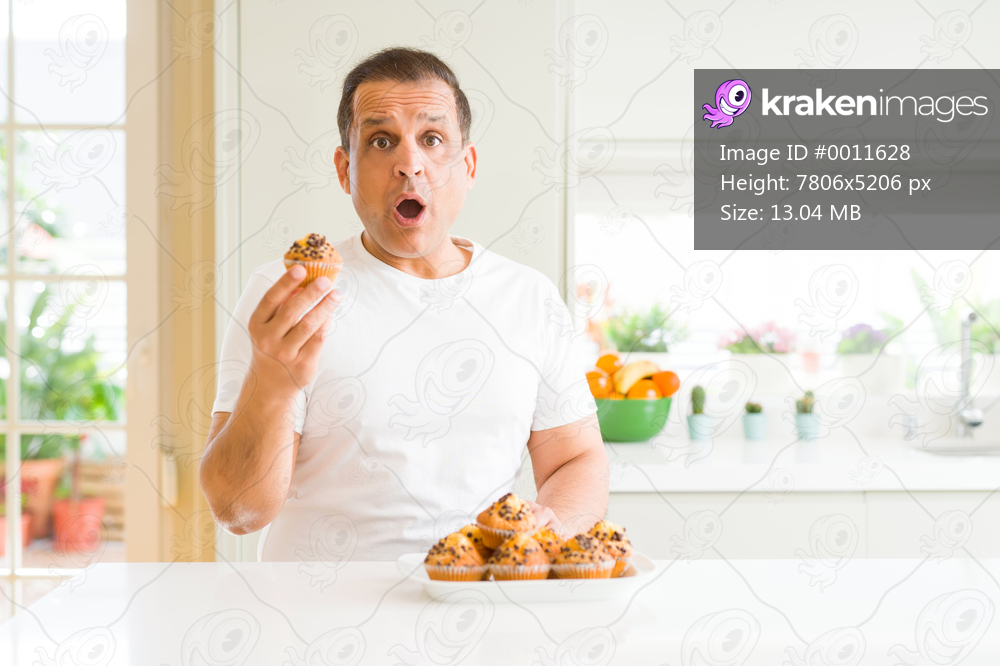 Middle age man eating chocolate chip muffins at home scared in shock with a surprise face, afraid and excited with fear expression