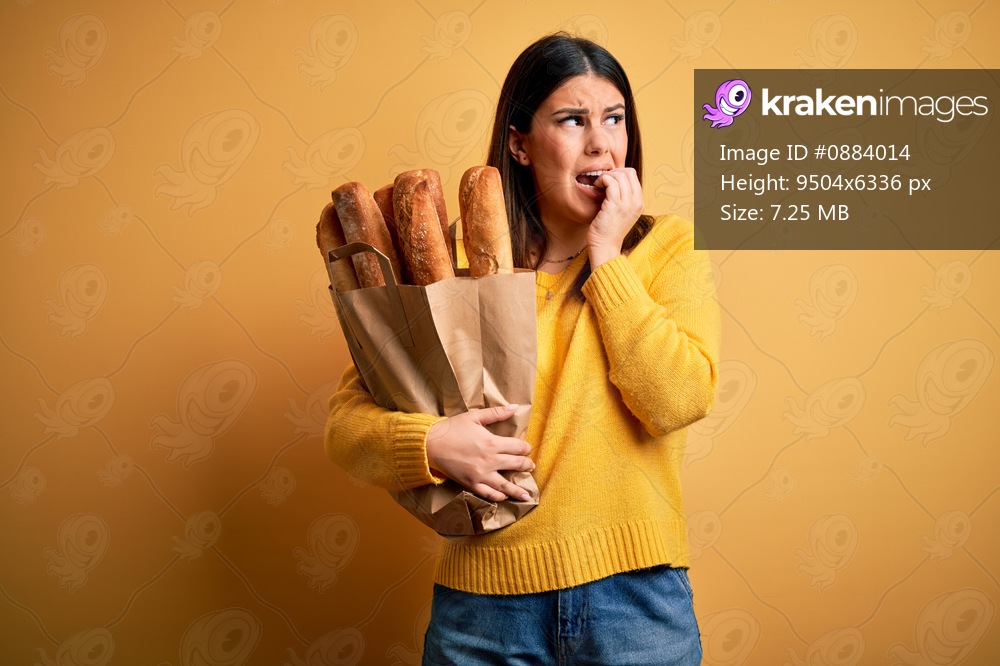 Young beautiful woman holding a bag of fresh healthy bread over yellow background looking stressed and nervous with hands on mouth biting nails. Anxiety problem.