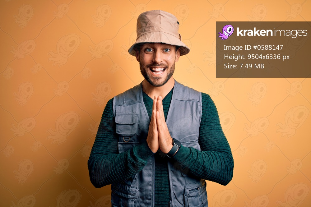 Handsome tourist man with beard on vacation wearing explorer hat over yellow background praying with hands together asking for forgiveness smiling confident.