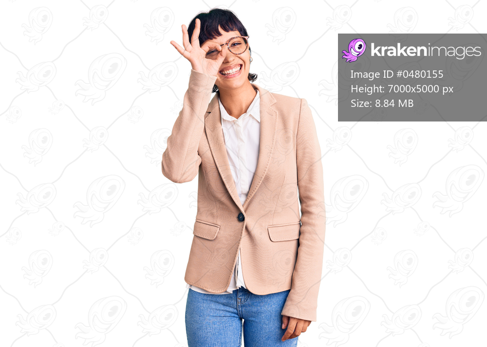 Young brunette woman with short hair wearing business jacket and glasses doing ok gesture with hand smiling, eye looking through fingers with happy face. 