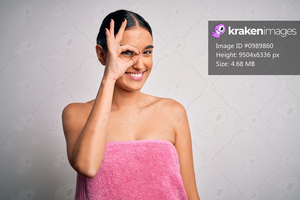 Young beautiful brunette woman wearing towel after shower over isolated white background doing ok gesture with hand smiling, eye looking through fingers with happy face.