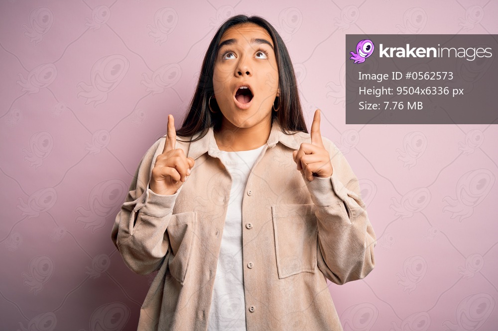 Young beautiful asian woman wearing casual shirt standing over pink background amazed and surprised looking up and pointing with fingers and raised arms.