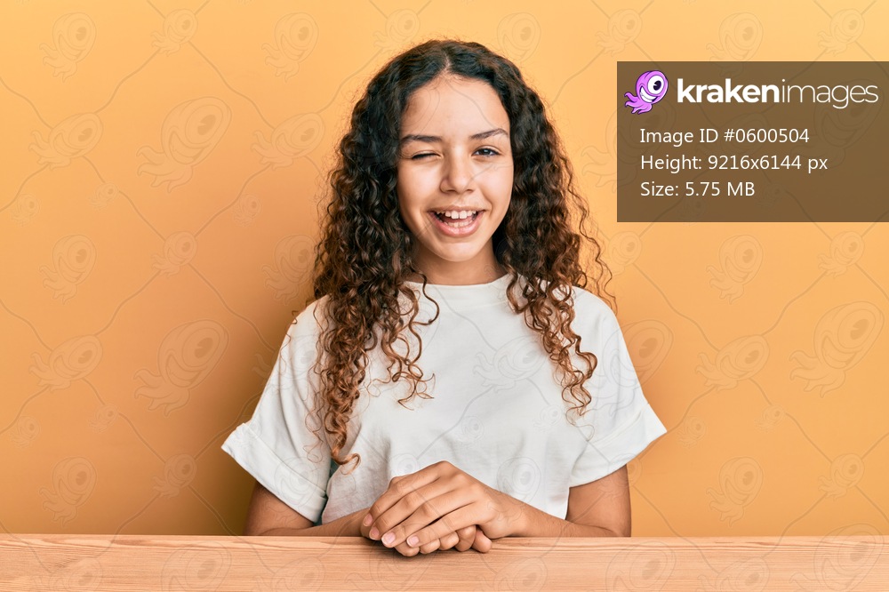 Teenager hispanic girl wearing casual clothes sitting on the table winking looking at the camera with sexy expression, cheerful and happy face. 