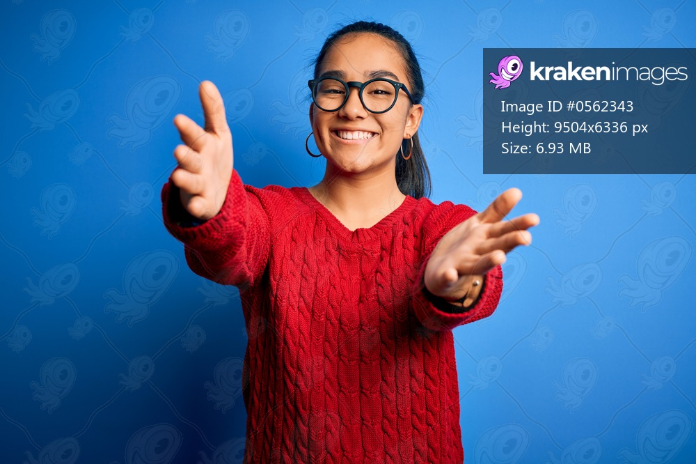 Young beautiful asian woman wearing casual sweater and glasses over blue background looking at the camera smiling with open arms for hug. Cheerful expression embracing happiness.