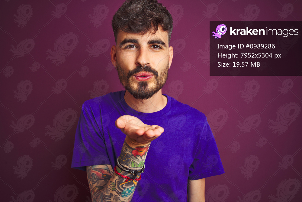 Young man with tattoo wearing t-shirt standing over isolated purple background looking at the camera blowing a kiss with hand on air being lovely and sexy. Love expression.