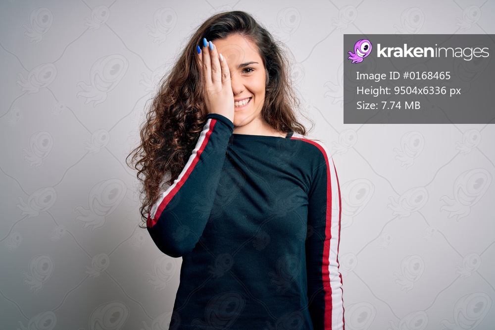 Young beautiful woman with curly hair wearing casual sweater over isolated white background covering one eye with hand, confident smile on face and surprise emotion.