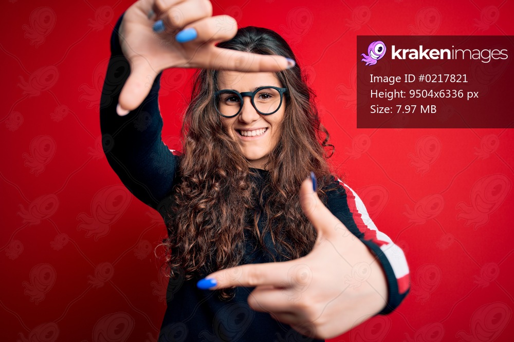 Young beautiful woman with curly hair wearing sweater and glasses over red background smiling making frame with hands and fingers with happy face. Creativity and photography concept.