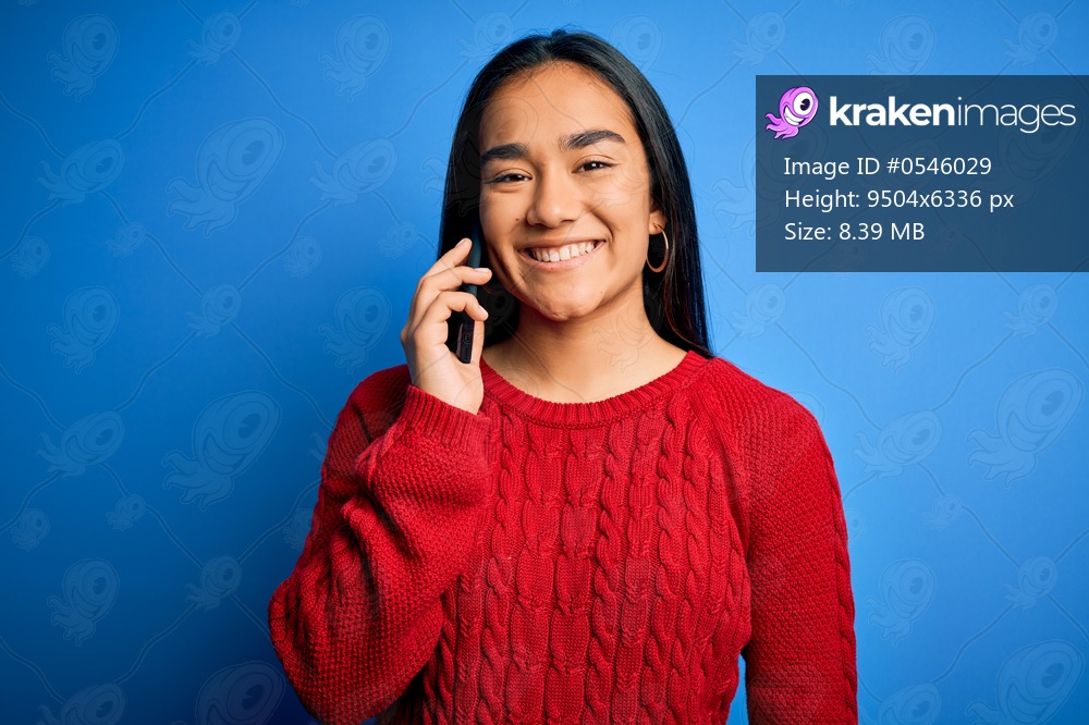 Young beautiful asian woman having conversation talking on smartphone over blue background with a happy face standing and smiling with a confident smile showing teeth