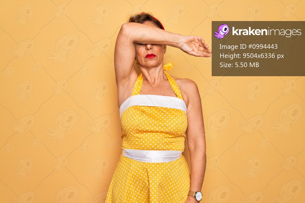 Middle age senior pin up woman wearing 50s style retro dress over yellow background covering eyes with arm, looking serious and sad. Sightless, hiding and rejection concept