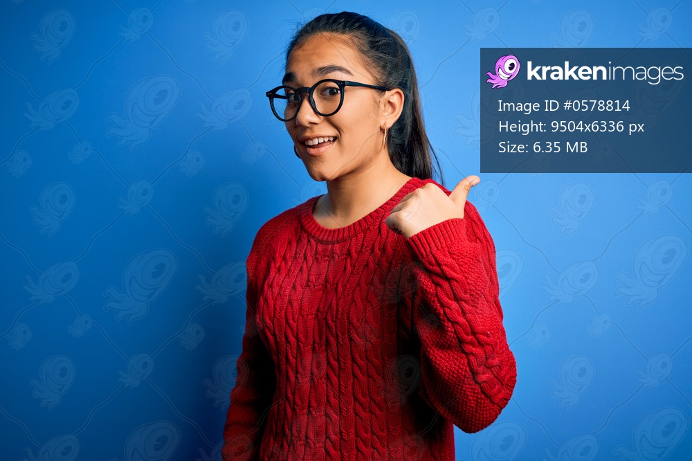 Young beautiful asian woman wearing casual sweater and glasses over blue background smiling with happy face looking and pointing to the side with thumb up.
