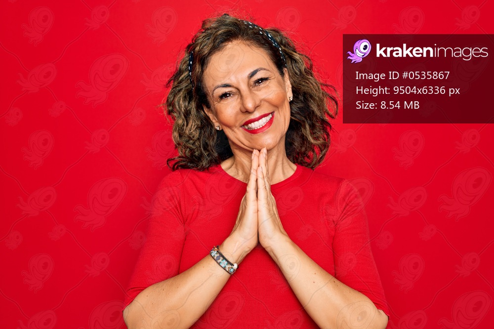 Middle age senior brunette woman wearing casual t-shirt standing over red background praying with hands together asking for forgiveness smiling confident.