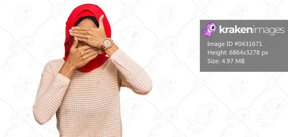 Young african american woman wearing traditional islamic hijab scarf covering eyes and mouth with hands, surprised and shocked. hiding emotion 