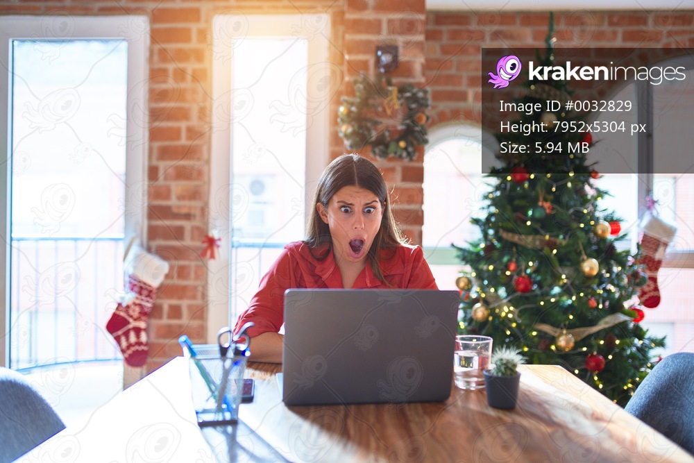 Beautiful woman sitting at the table working with laptop at home around christmas tree afraid and shocked with surprise and amazed expression, fear and excited face.