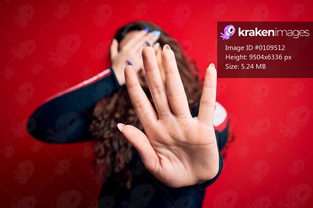 Young beautiful woman with curly hair wearing sweater and glasses over red background covering eyes with hands and doing stop gesture with sad and fear expression. Embarrassed and negative concept.