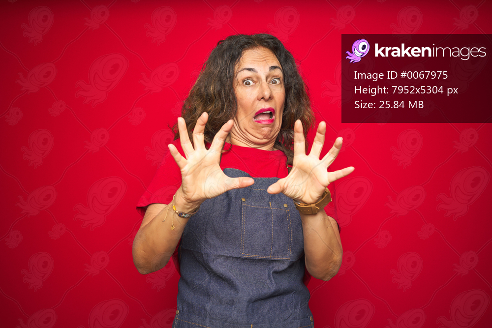 Middle age senior woman wearing apron uniform over red isolated background afraid and terrified with fear expression stop gesture with hands, shouting in shock. Panic concept.