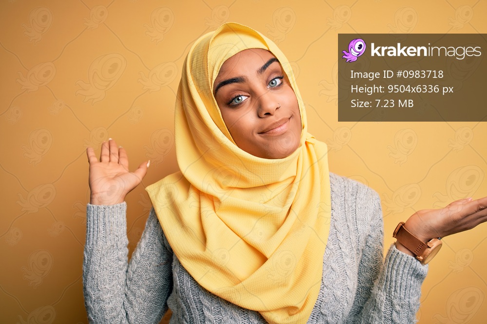 Young beautiful african american girl wearing muslim hijab over isolated yellow background clueless and confused expression with arms and hands raised. Doubt concept.