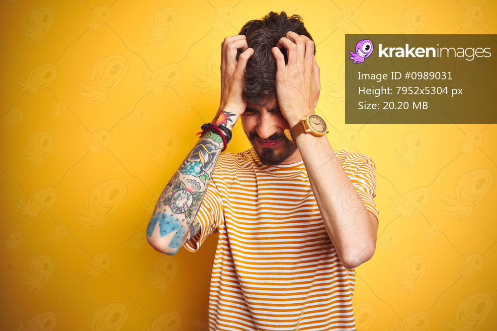 Young man with tattoo wearing striped t-shirt standing over isolated yellow background suffering from headache desperate and stressed because pain and migraine. Hands on head.