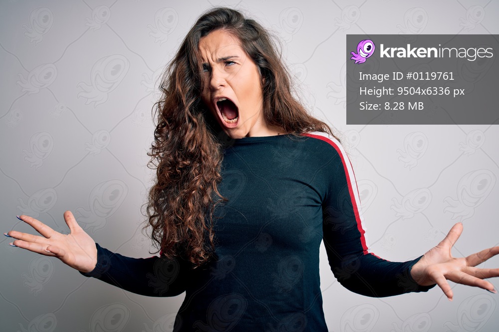 Young beautiful woman with curly hair wearing casual sweater over isolated white background crazy and mad shouting and yelling with aggressive expression and arms raised. Frustration concept.