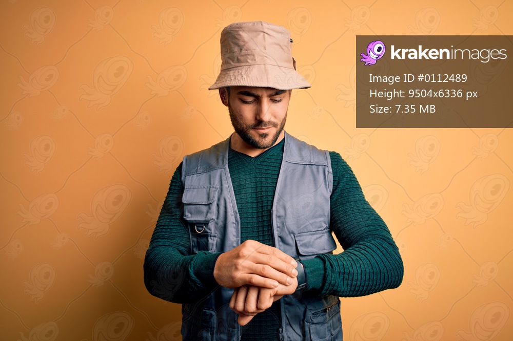 Handsome tourist man with beard on vacation wearing explorer hat over yellow background Checking the time on wrist watch, relaxed and confident