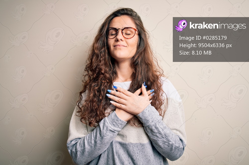 Young beautiful woman with curly hair wearing sweater and glasses over white background smiling with hands on chest with closed eyes and grateful gesture on face. Health concept.