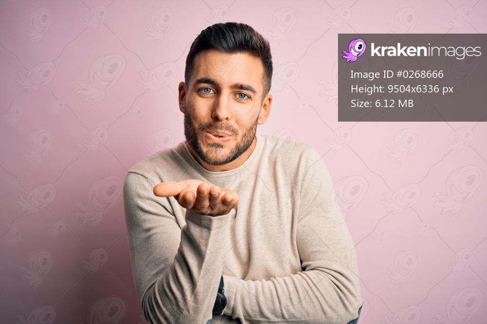 Young handsome man wearing casual sweater standing over isolated pink background looking at the camera blowing a kiss with hand on air being lovely and sexy. Love expression.