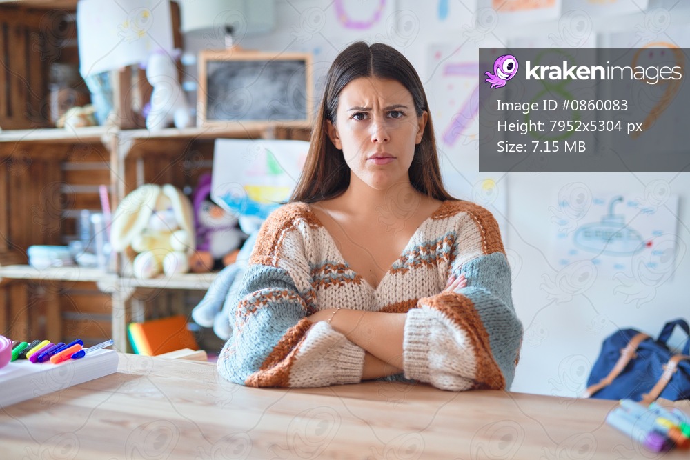 Young beautiful teacher woman wearing sweater and glasses sitting on desk at kindergarten skeptic and nervous, disapproving expression on face with crossed arms. Negative person.