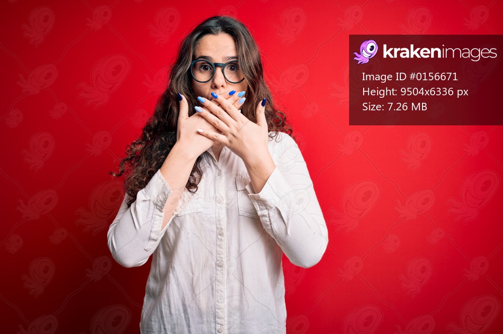 Young beautiful woman with curly hair wearing shirt and glasses over red background shocked covering mouth with hands for mistake. Secret concept.