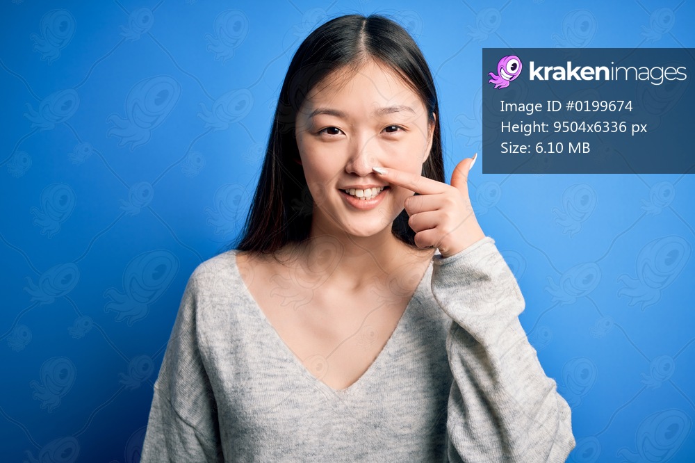 Young beautiful asian woman wearing casual sweater standing over blue isolated background Pointing with hand finger to face and nose, smiling cheerful. Beauty concept