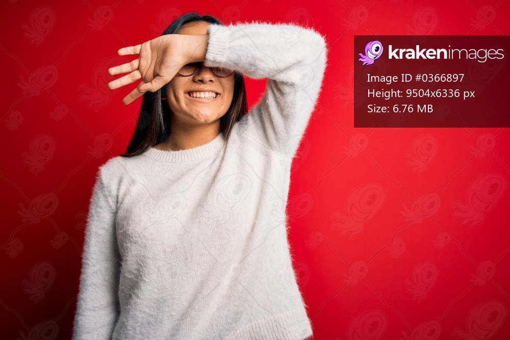 Young beautiful asian woman wearing casual sweater and glasses over red background covering eyes with arm smiling cheerful and funny. Blind concept.