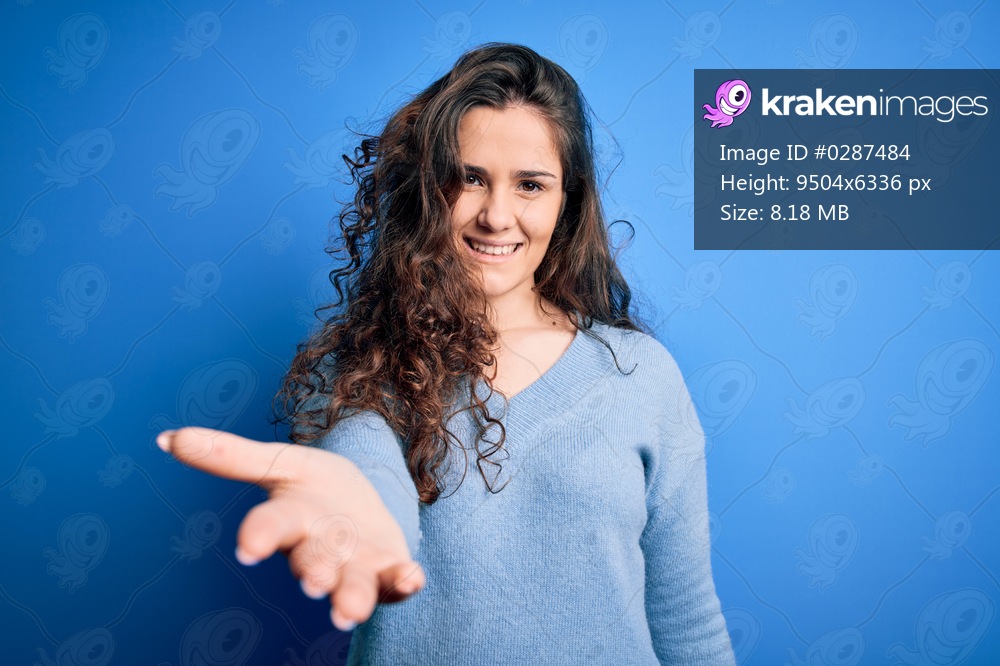 Young beautiful woman with curly hair wearing blue casual sweater over isolated background smiling cheerful offering palm hand giving assistance and acceptance.
