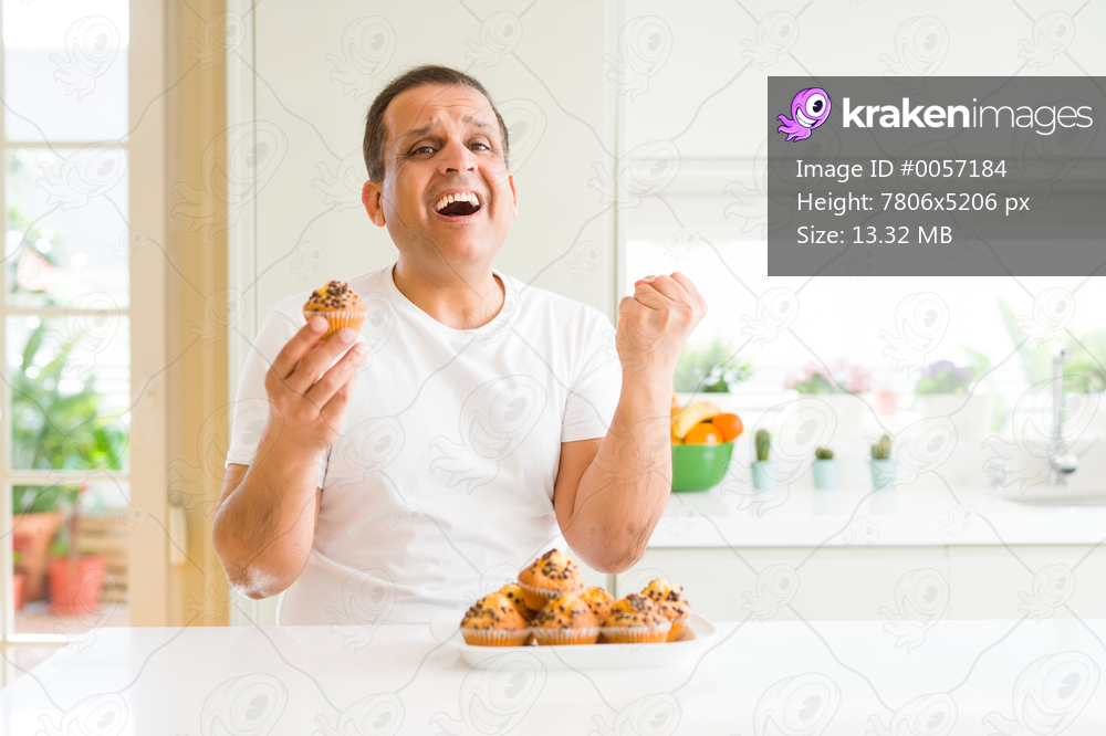 Middle age man eating chocolate chip muffins at home screaming proud and celebrating victory and success very excited, cheering emotion