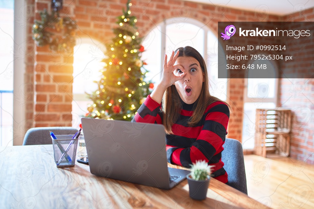Beautiful woman sitting at the table working with laptop at home around christmas tree doing ok gesture shocked with surprised face, eye looking through fingers. Unbelieving expression.