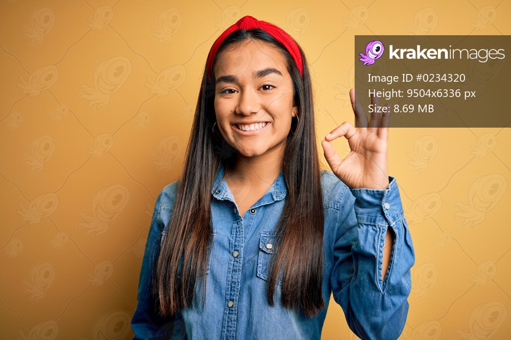 Young beautiful asian woman wearing casual denim shirt and diadem over yellow background smiling positive doing ok sign with hand and fingers. Successful expression.