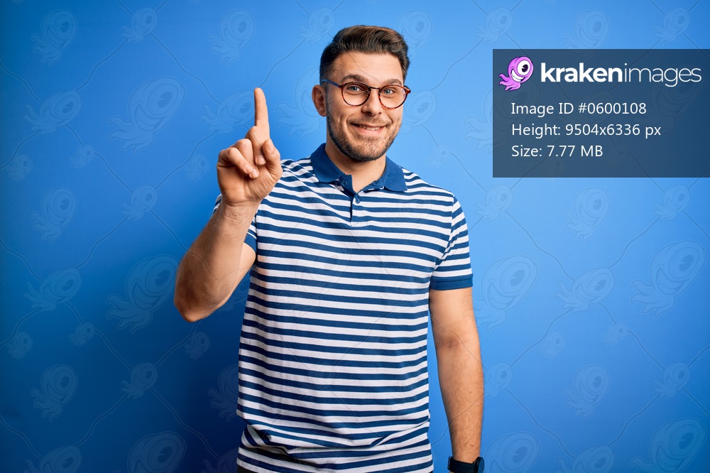 Young man with blue eyes wearing glasses and casual striped t-shirt over blue background showing and pointing up with finger number one while smiling confident and happy.