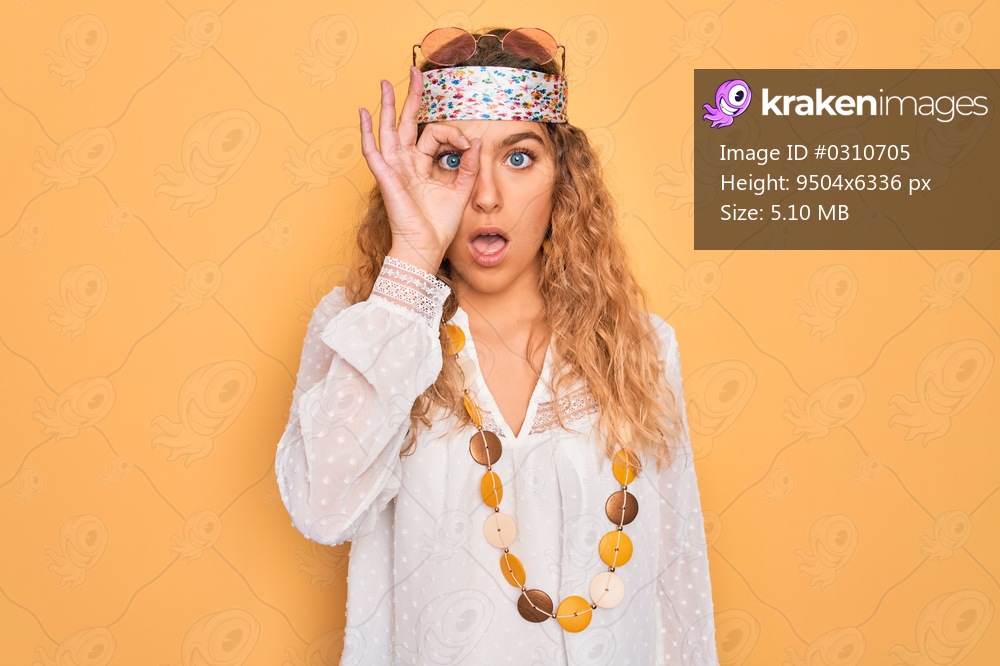 Young beautiful blonde hippie woman with blue eyes wearing sunglasses and accessories doing ok gesture shocked with surprised face, eye looking through fingers. Unbelieving expression.