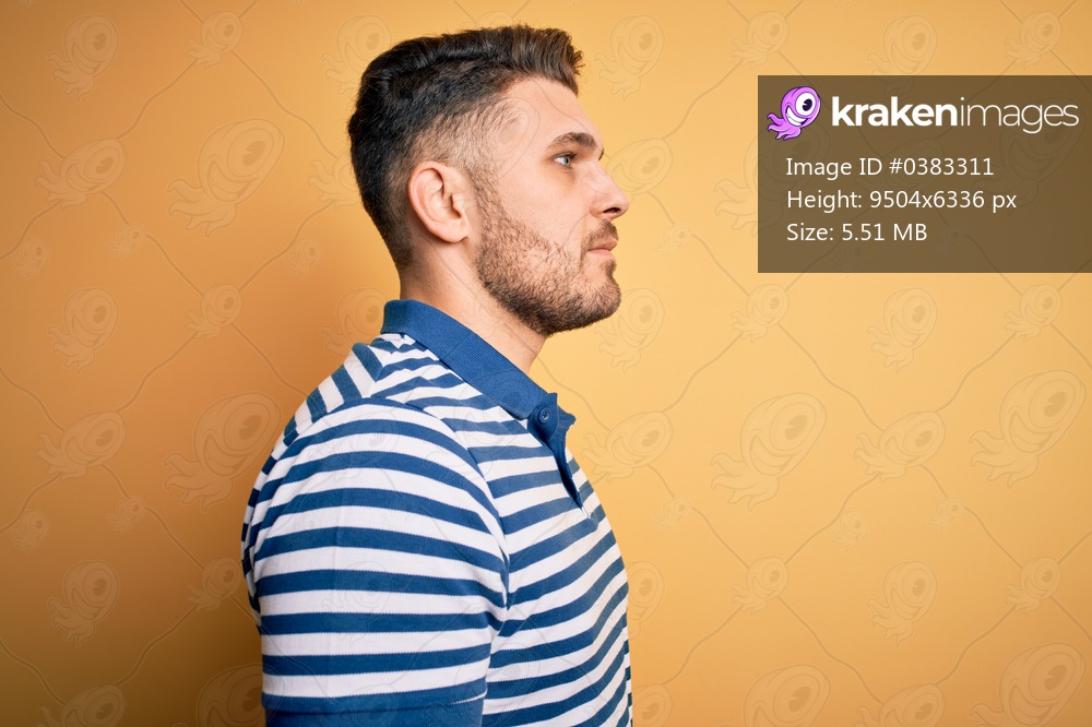 Young man with blue eyes wearing casual striped t-shirt over yellow background looking to side, relax profile pose with natural face with confident smile.