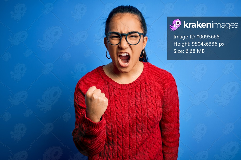 Young beautiful asian woman wearing casual sweater and glasses over blue background angry and mad raising fist frustrated and furious while shouting with anger. Rage and aggressive concept.